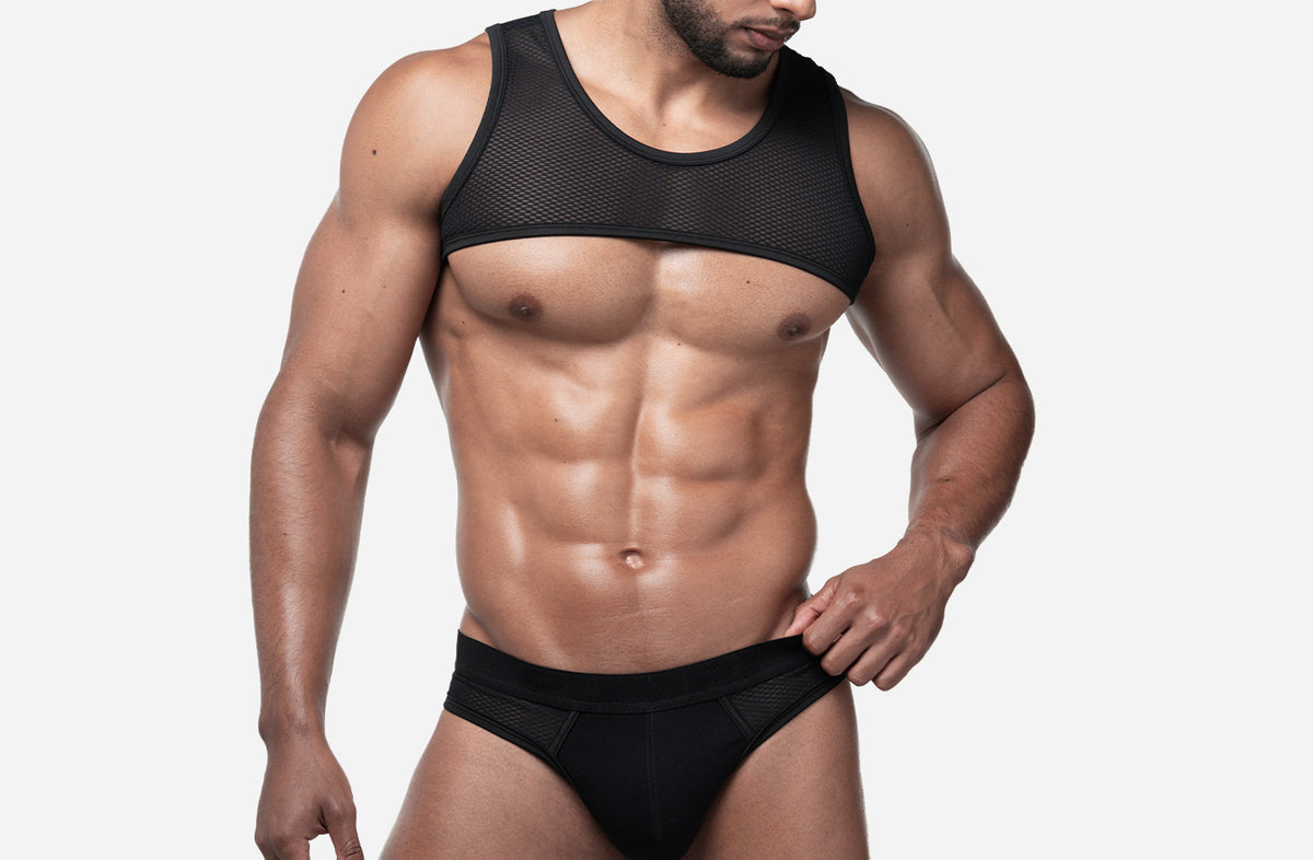 A muscular man wearing a black mesh PUMP harness and black briefs, shown from the waist up against a light gray studio background.