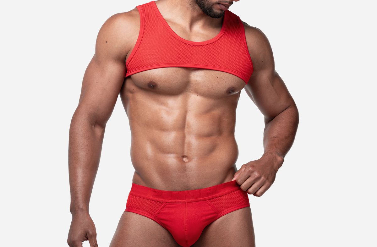 A muscular man wearing a matching red two-piece underwear set consisting of a sleeveless tank top harness and briefs, shown against a plain light gray background.  The brand is PUMP jockstraps and harnesses.